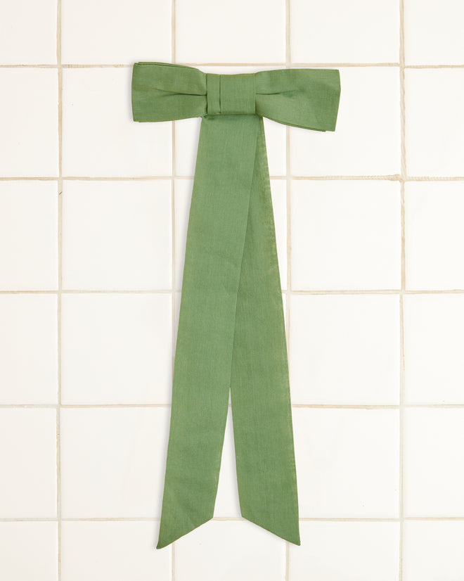 The Bow Barrette - Green, crafted from cotton voile with long tails, is elegantly hanging on a white tiled wall.