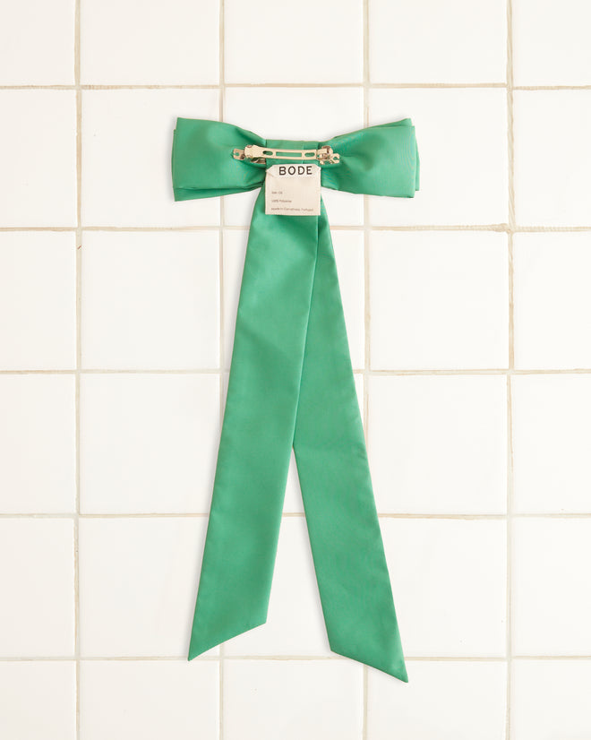 A Harbor Bow Barrette in green, featuring long tails and made from recycled polyester, is affixed to a tiled wall. A small label marked 