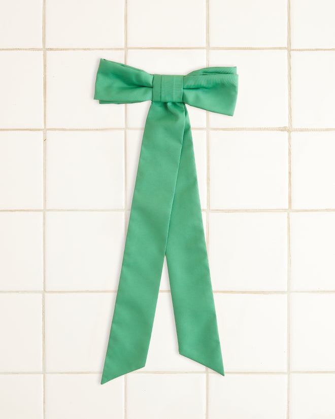 The Harbor Bow Barrette - Green, featuring a green ribbon bow with two long tails and crafted from recycled polyester, is displayed against a light beige background.