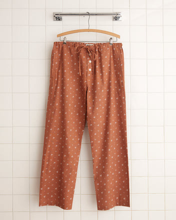 The Harbor Tumble Pajama Pants, featuring a white leaf pattern on brown fabric, button details, and a drawstring, made in the USA, are hanging on a wooden hanger against a white tiled wall.