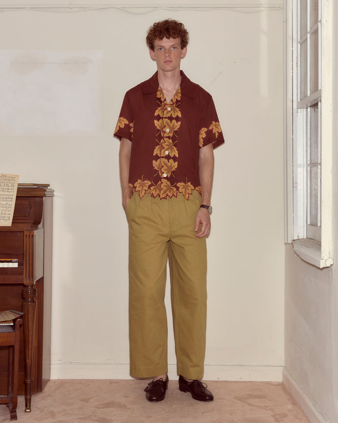 A man in a Harvest Floor Short Sleeve Shirt and tan pants stands indoors by a piano, hands in pockets, facing the camera.
