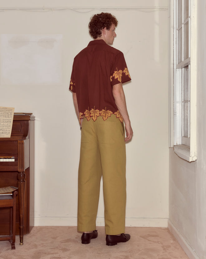 A man in a brown Harvest Floor Short Sleeve Shirt with yellow leaf patterns and khaki pants stands facing the corner of a room.