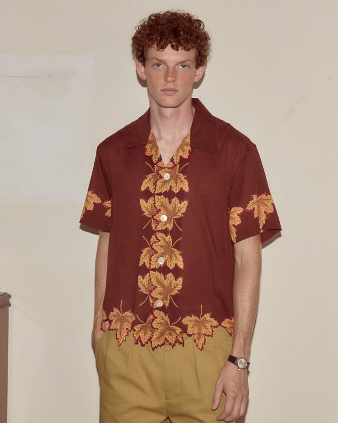 A man with curly hair stands indoors wearing a Harvest Floor Short Sleeve Shirt featuring yellow leaf designs and tan pants.