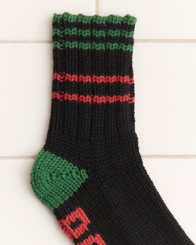 The Hawthorne Stripe Socks, hand-knit in black and green with red stripes, crafted from cozy merino wool, are laid out on a white tiled surface.