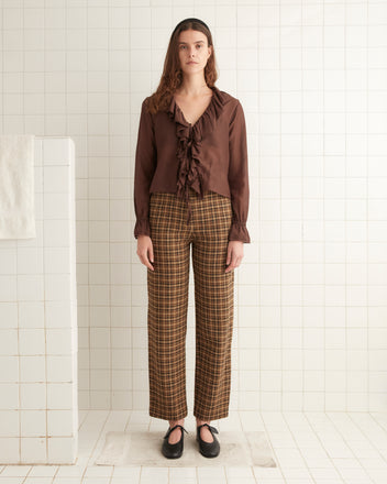 A woman stands in a tiled room wearing the Heartwood Flounce Blouse, a lightweight cotton/silk blend with ruffled design and tie-front closures, paired with plaid pants. She has long brown hair and black shoes, and she looks directly at the camera.