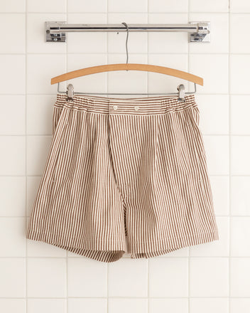 The Heathway Stripe Boxers - S, featuring beige and white stripes and crafted from deadstock fabric, are elegantly displayed on a wooden hanger against a tiled wall.