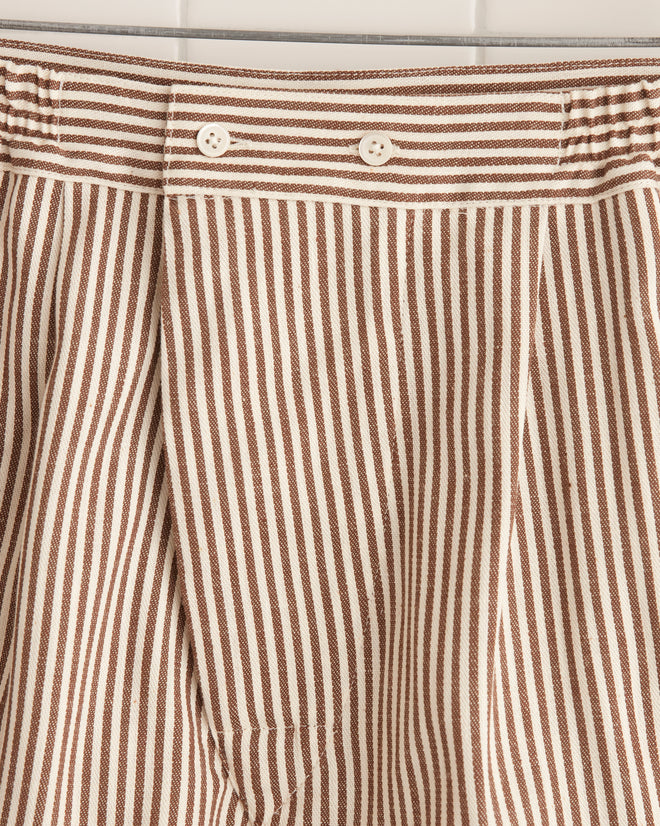 The Heathway Stripe Boxers - S, featuring beige and white stripes and crafted from deadstock fabric, are elegantly displayed on a wooden hanger against a tiled wall.