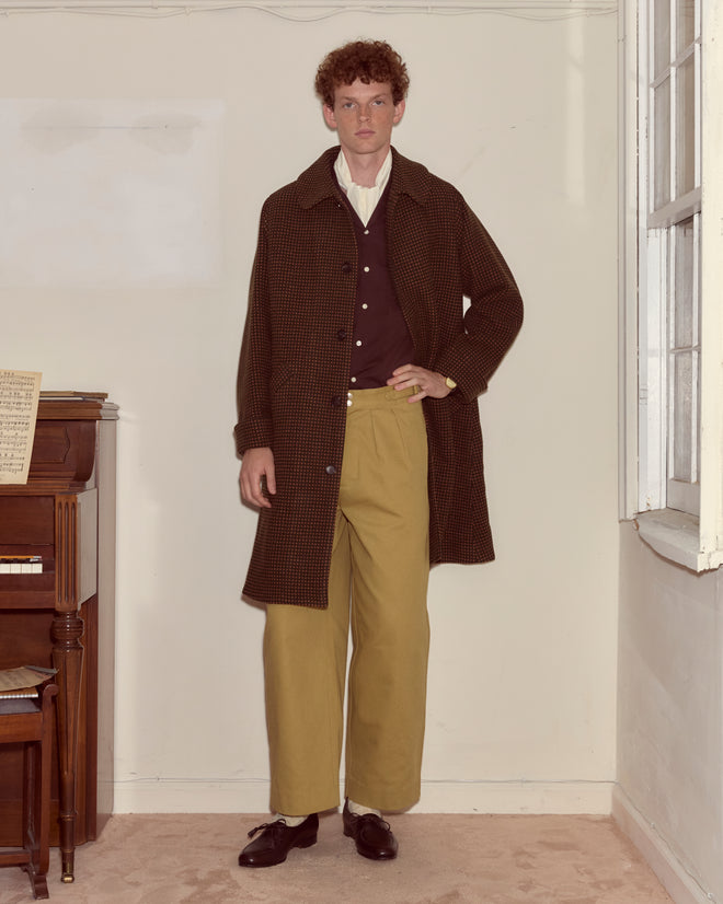 A man wearing the Hedgerow Coat - Green, a heavyweight wool overcoat with a swing silhouette, paired with tan pants and loafers, stands indoors beside a piano and a window.