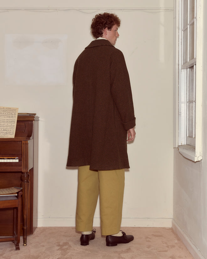 A person in the Hedgerow Coat - Green and tan pants stands facing a corner near a piano and a window.