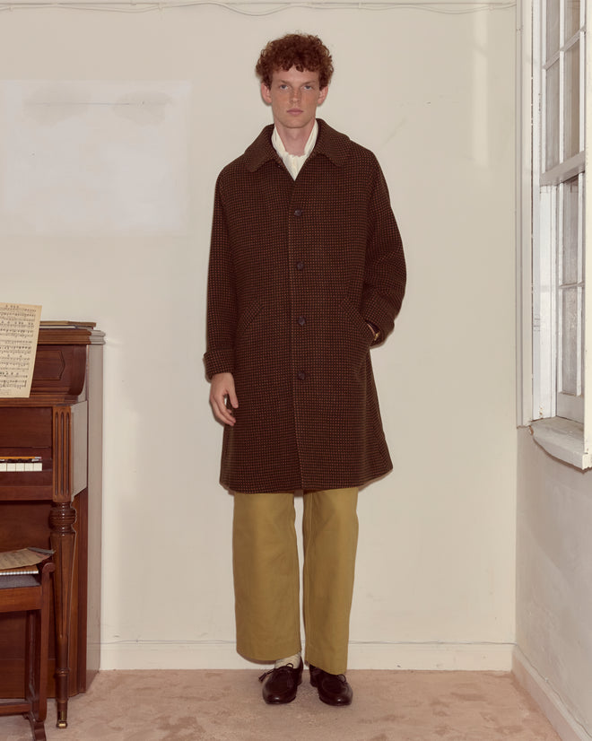 A man in a Hedgerow Coat - Green and tan pants stands indoors beside a piano near a window.