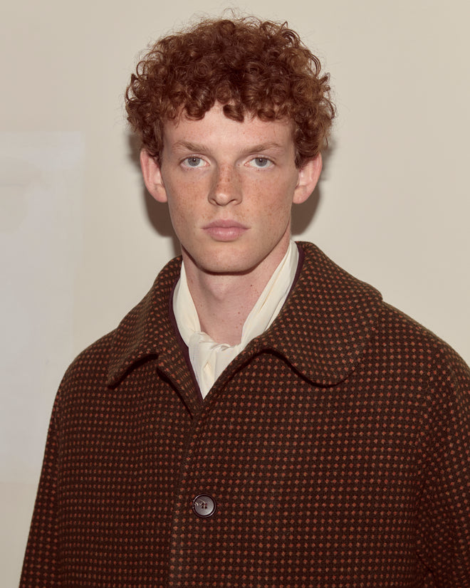 A young man with curly red hair wears the Hedgerow Coat in deep green, a heavyweight wool overcoat with a swing silhouette, paired with a cream scarf, standing against a plain background.