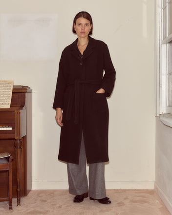 A woman stands indoors by a piano wearing the Hemlock Coat, a long black wool coat with a belted waist, paired with gray pants and black shoes.