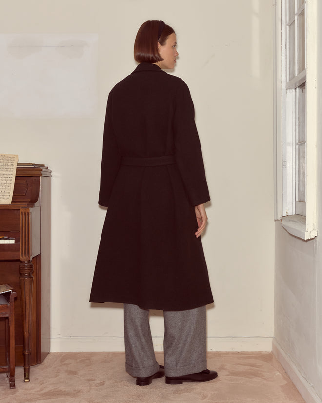 A woman wearing the black Hemlock Coat with a belted waist and gray pants stands by a piano, gazing out a sunlit window in a minimalist room.