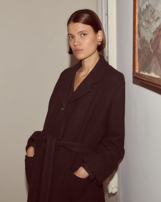 A woman with short brown hair stands indoors by a light wall and framed painting, wearing the Hemlock Coat—a black wool coat with a belted waist.