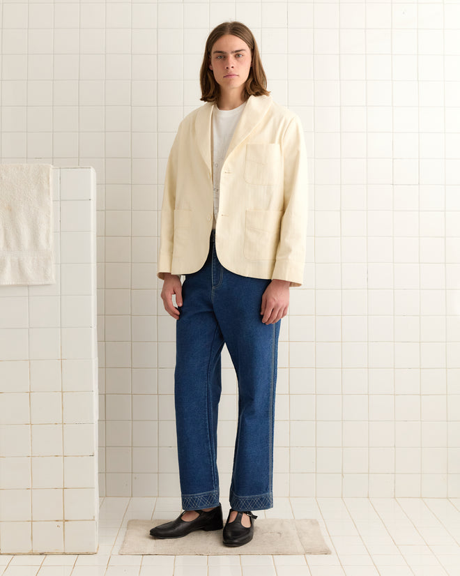 The individual is dressed in a Herringbone Smoking Jacket with a shawl collar over a white shirt, paired with blue jeans and black shoes. They're standing in a tiled room, the cuffed sleeves subtly highlighting the elegance of the cream herringbone twill fabric.