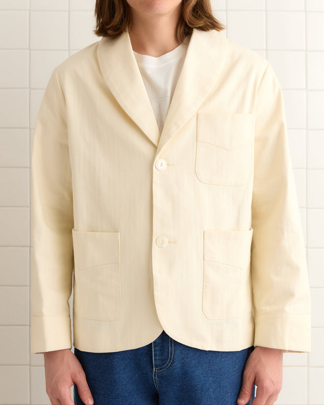 A person in a Herringbone Smoking Jacket with cuffed sleeves and pocket details, wearing blue jeans and standing in front of a tiled wall.