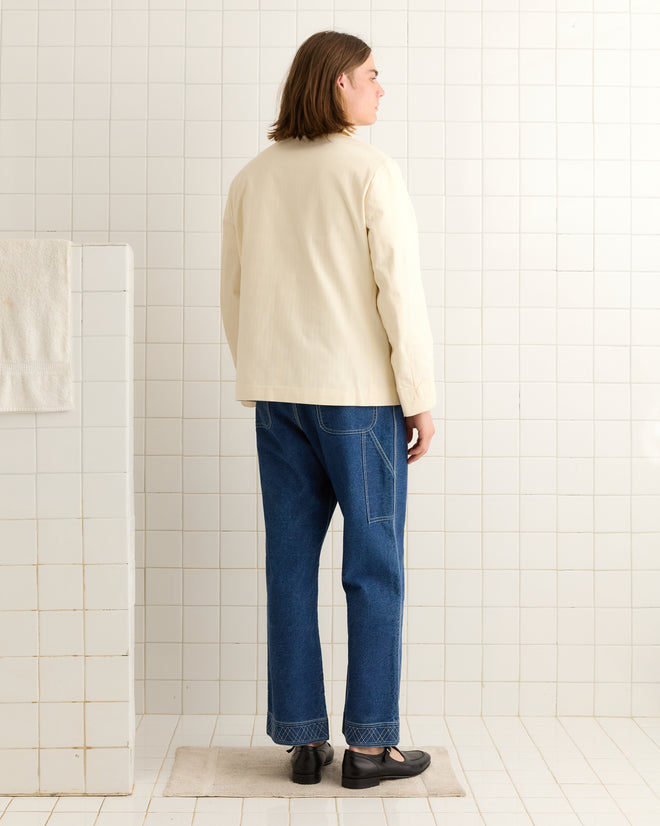 A person with long hair in a cream Herringbone Smoking Jacket and blue jeans stands on a tiled floor, facing a tiled wall.