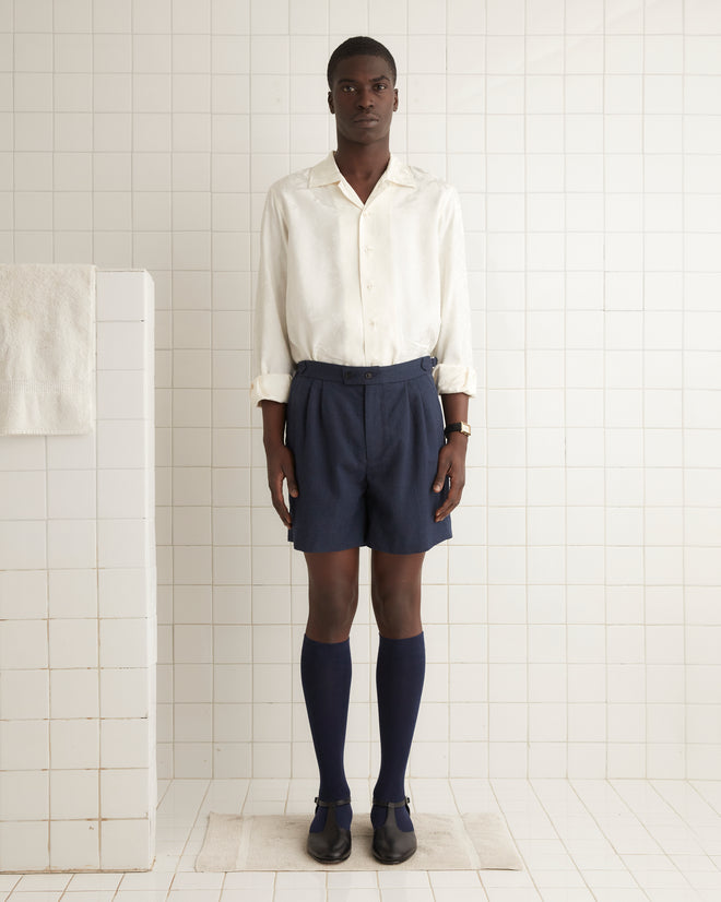 A person in a plaid blazer, white shirt, and Herringbone Wool Shorts - 32 with side-buckle waist adjusters stands against a wooden background.