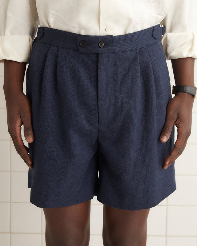 A person in a plaid blazer, white shirt, and Herringbone Wool Shorts - 32 with side-buckle waist adjusters stands against a wooden background.