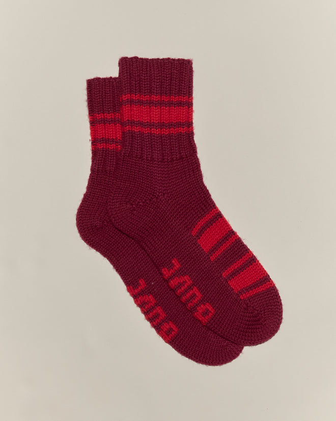 A pair of Herrington Stripe Socks - Burgundy, hand-knit in maroon with red stripes and "DUDE" on the soles, shown on a light background.