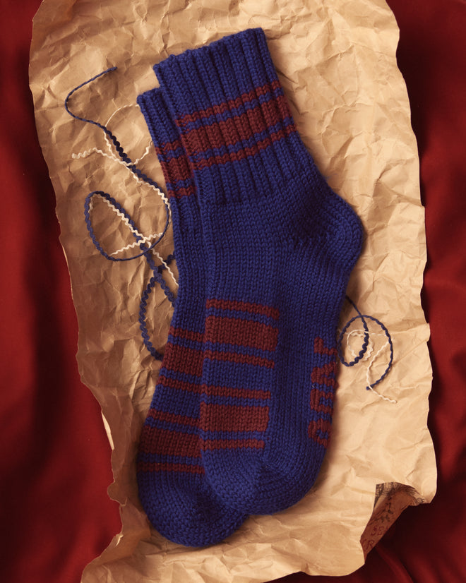 Two pairs of Herrington Stripe Socks - Blue, crafted from soft merino wool, rest on crumpled paper with a string nearby, all set against a rich red fabric background.