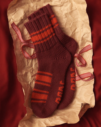 The Herrington Stripe Socks in Burgundy, featuring a cozy merino wool blend and red and orange stripes, are beautifully displayed on crumpled paper with red ribbon accents.