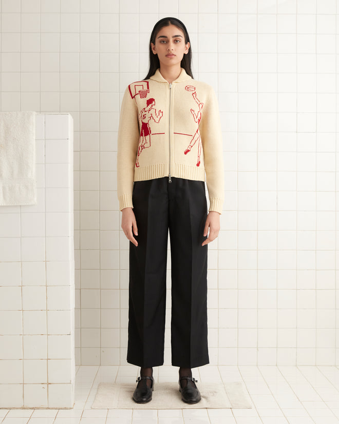 A person wearing a Hoop Cardigan in cream knit with red designs and black pants is standing in a tiled room.