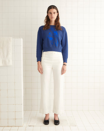 A person stands in a white-tiled room wearing a blue cotton Hoop Sweatshirt with a graphic and white pants, paired with black shoes.