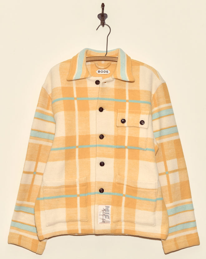 The Horizon Plaid Blanket Jacket - XS/S is a unique yellow plaid wool jacket with turquoise and white stripes, black buttons, and large front pockets, made in New York. It's pictured hanging on a hook.