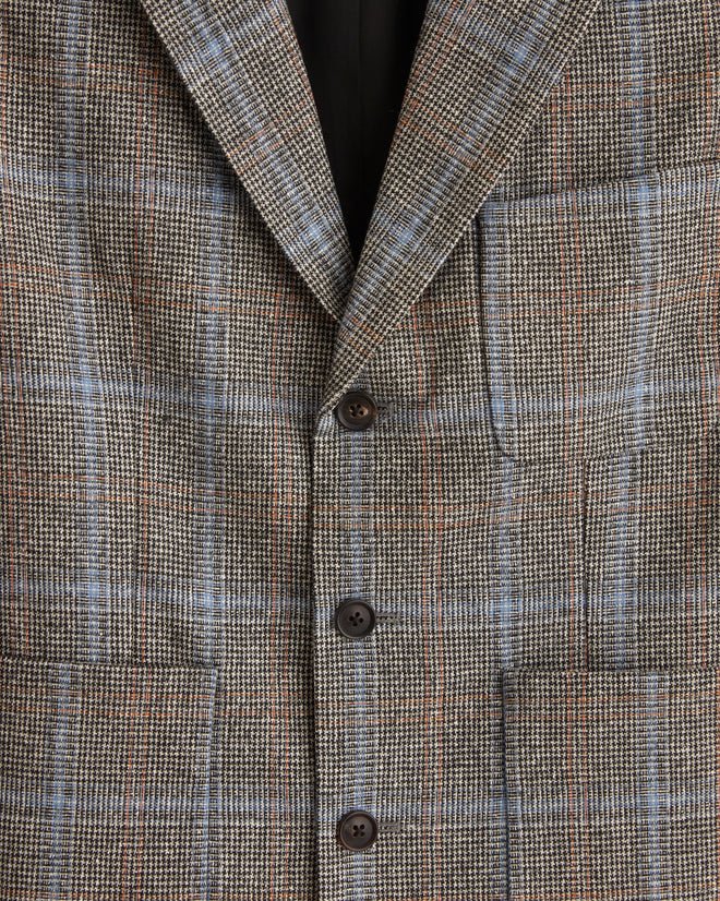 The Houndstooth Pigment Suit Jacket - 38, made from pure wool, hangs elegantly on a wall-mounted hanger against a white tiled background, evoking the classic style of 1960s suiting fabric.