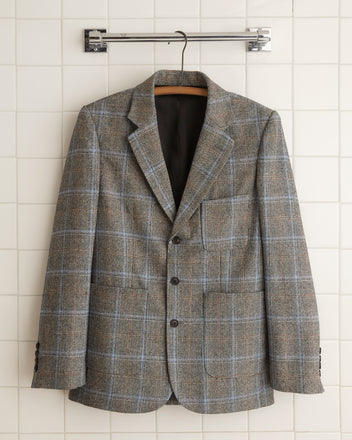 The Houndstooth Pigment Suit Jacket - 38, made from pure wool, hangs elegantly on a wall-mounted hanger against a white tiled background, evoking the classic style of 1960s suiting fabric.