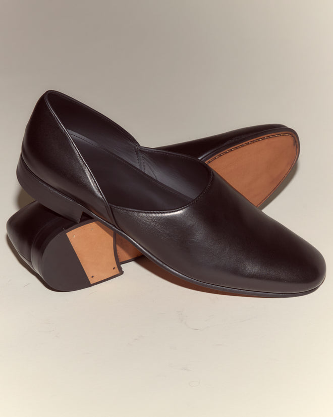 A pair of Leather House Shoes - Black, crafted in Italy from black Napa leather with brown soles, one shoe resting atop the other.