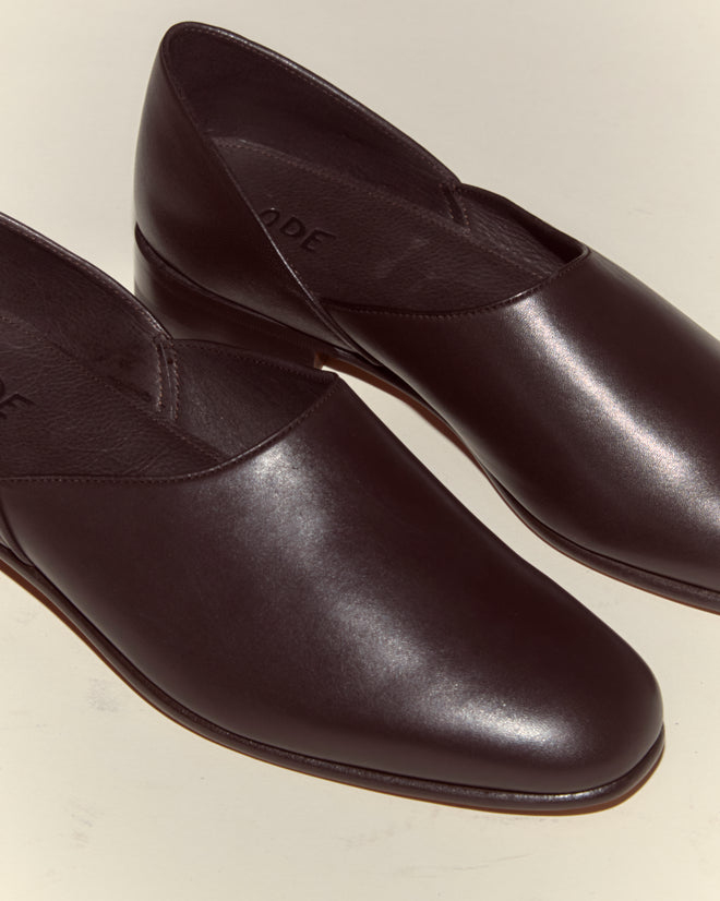 A pair of Leather House Shoes - Brown, featuring sleek dark brown Napa leather and a minimalist slip-on design, is displayed on a light surface.
