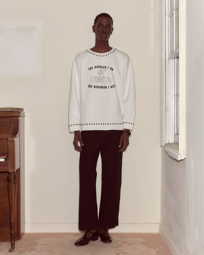 A person stands indoors by a window and small table, wearing the Hurrier Sweatshirt with striped black and white trim and black pants.