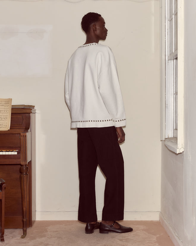 A person stands indoors by a window, wearing a Hurrier Sweatshirt with striped black and white trim, paired with black pants and black shoes.