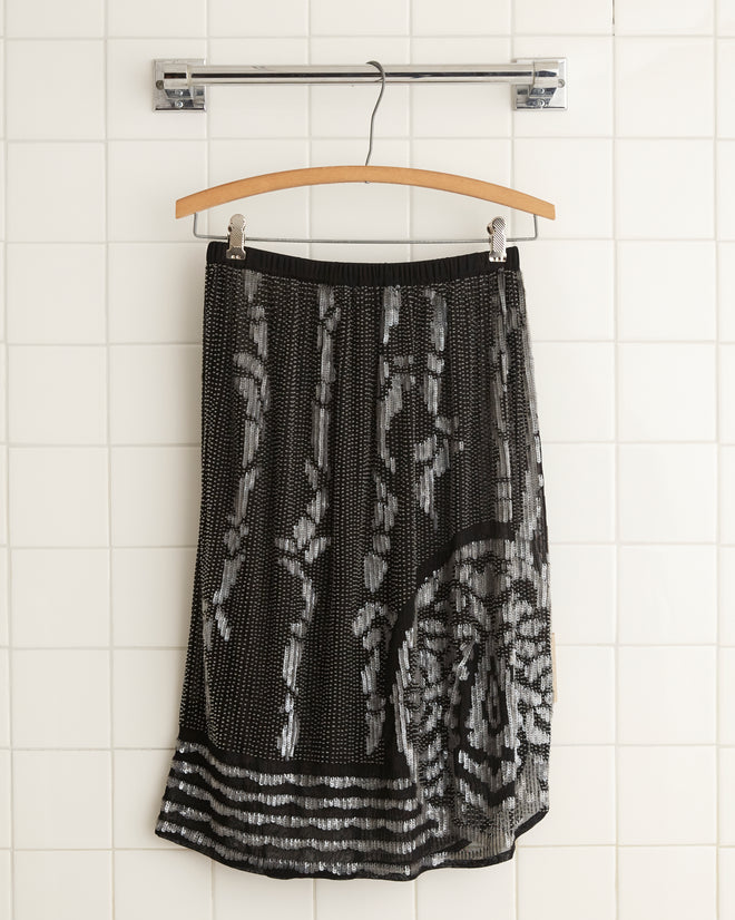 The Hyatt Skirt - Black/Multi, featuring a white abstract pattern and an asymmetrical scalloped hem, hangs on a wooden hanger against a white tiled wall.