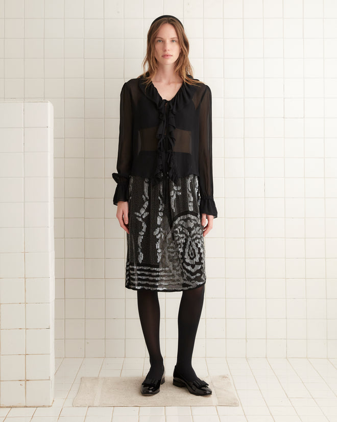 The Hyatt Skirt - Black/Multi, featuring a white abstract pattern and an asymmetrical scalloped hem, hangs on a wooden hanger against a white tiled wall.