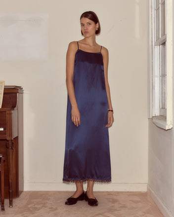 A woman in the unique Hypnos Dress made from deadstock satin and black flats stands indoors by a piano and a window.