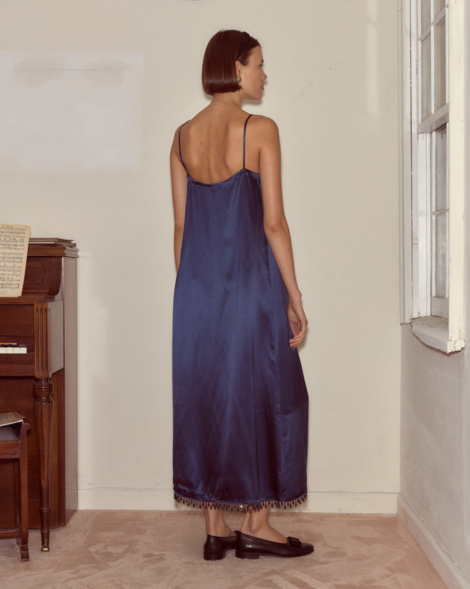 A woman wearing the Hypnos Dress made of deadstock satin stands indoors, facing away, near a piano and a large window.