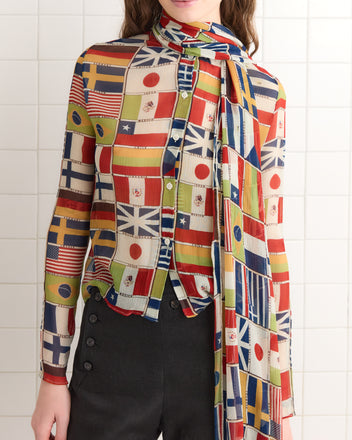 A person stands against a white-tiled background, wearing the International Blouse made of silk crepe with a vivid flag print, paired with dark pants.