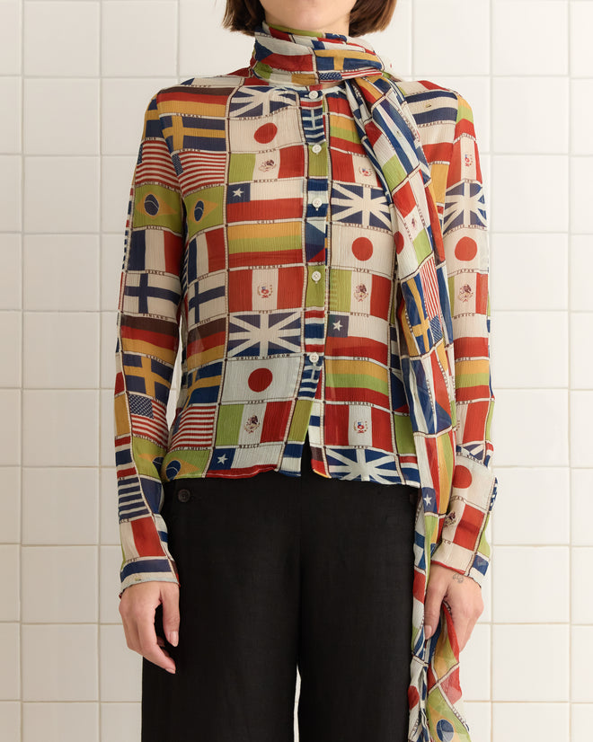 A person wearing an International Blouse, a vibrant shirt adorned with various international flags, stands against a tiled wall.