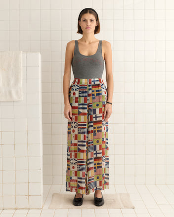 A person dressed in a gray tank top and International Pants, with a colorful, vibrant flag pattern, stands on a bath mat in a tiled room.