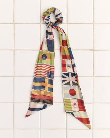 The International Silk Scrunchie, featuring long ribbons and a vibrant flag print, elegantly hangs on a tiled wall—an eye-catching accessory for any occasion.