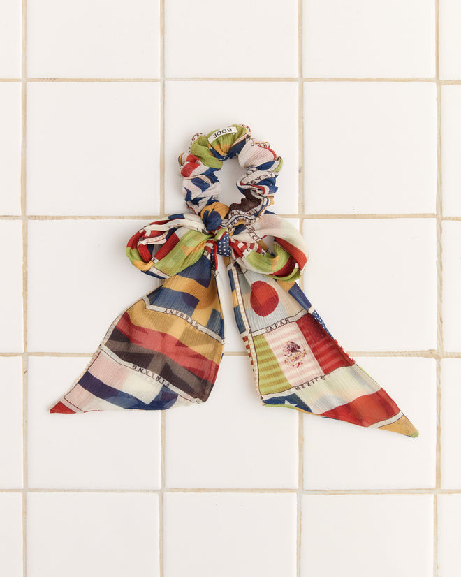 The International Silk Scrunchie, showcasing a vibrant flag print with ribbons, is beautifully crafted from silk and displayed on a crisp white tiled background—making it a stunning accessory for any collection.