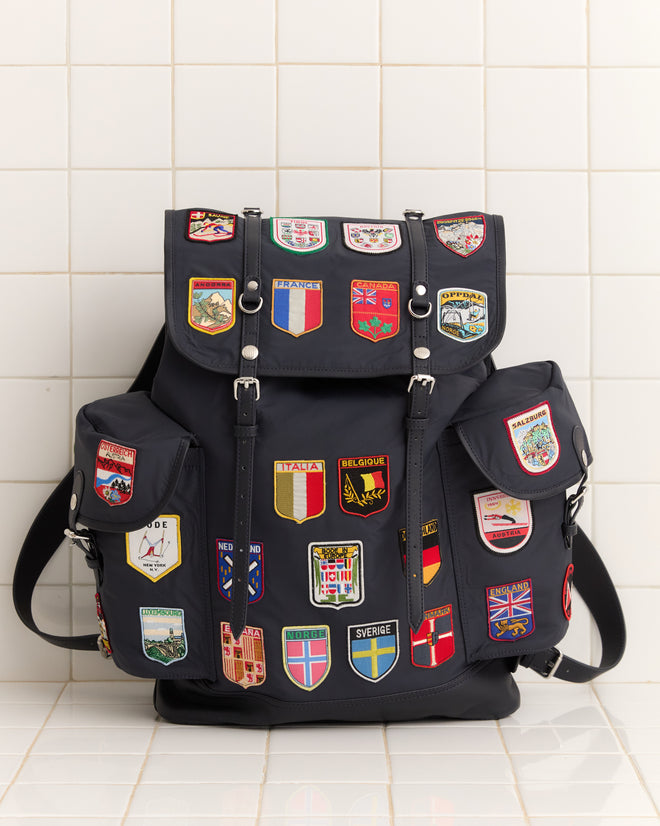 The Interrail Rucksack, featuring colorful country and city patches on black fabric, stands out against a white tiled wall for a vibrant accessory display.