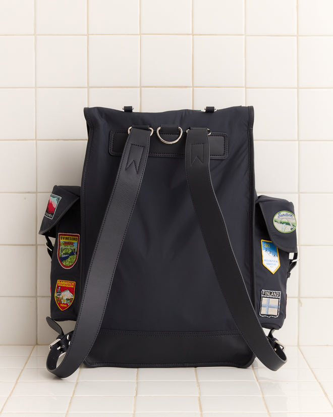 The Interrail Rucksack, featuring black fabric and side pocket patches, sits against a white tiled wall—ideal for carrying your favorite accessories.