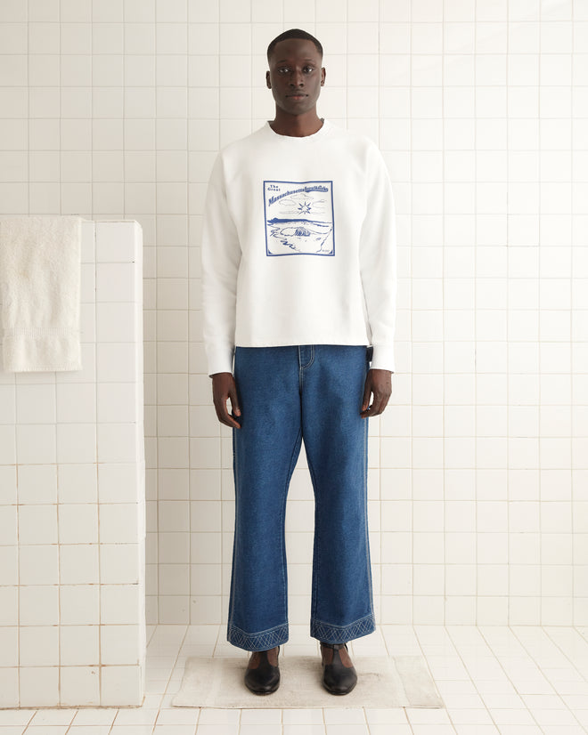 In a tiled room, an individual is wearing the Ironworks Crewneck—a heavyweight sweater featuring a blue graphic—paired with blue jeans. The cotton crewneck, crafted in Portugal, combines comfort and style, and a white towel hangs on a nearby rack.