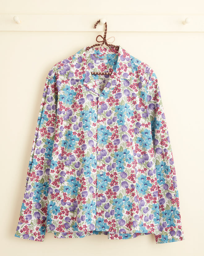 The Island Leiflower Shirt - L/XL, a long-sleeve button-up shirt crafted from midcentury deadstock fabric with a vibrant floral pattern featuring blue, purple, and red flowers, hangs on a hook against a white wall.