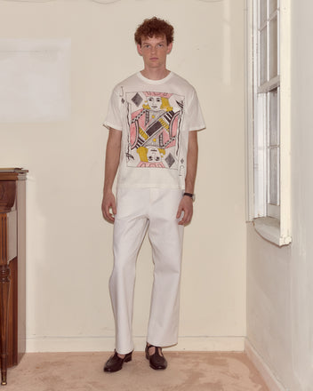 A man in white pants and a cream Jack Of Diamonds Pocket Tee with a bold card motif stands by a window, evoking an Alice Through the Looking Glass vibe.