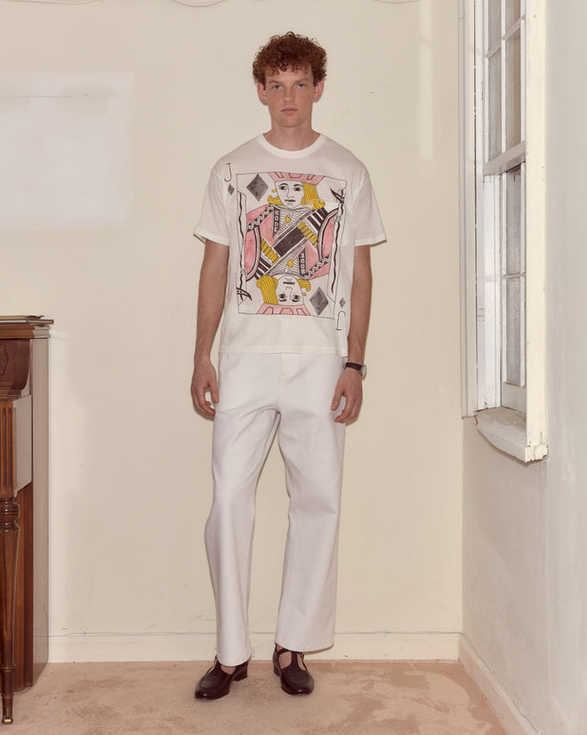 A man in white pants and a cream Jack Of Diamonds Pocket Tee with a bold card motif stands by a window, evoking an Alice Through the Looking Glass vibe.